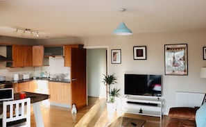 A modern kitchen with wooden cabinets, a dining area with a white chair, a TV, and a sofa.