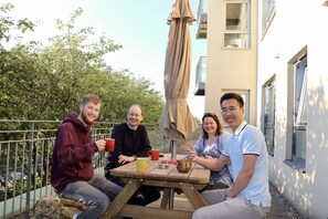 A group of people sitting at an outdoor wooden table with drinks, surrounded by a balcony with a metal railing and trees in the background.