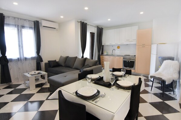 A modern living room with a dining area, a grey sofa, a white table set for four, and a kitchen in the background.