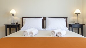 A neatly made bed with white linens and two rolled towels, flanked by two bedside tables with lamps and a cup on one.
