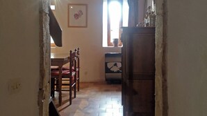Ein rundbogiger Durchgang führt in einen Raum mit Fliesenboden, einem Holztisch mit Stühlen, einem Fenster mit Vorhängen und einem Schrank.