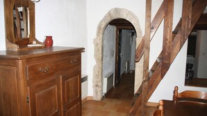 Eine hölzerne Kommode mit Schubladen, ein Spiegel und eine rote Kanne. Ein Steinenbogen führt zu einer Treppe.