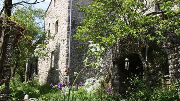 Ein Steinhaus mit einem Turm, umgeben von Grünpflanzen und blühenden Blumen.