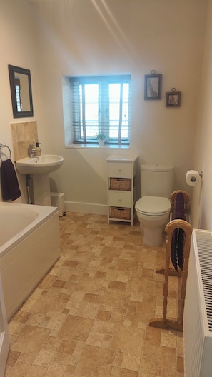 A bathroom with a sink, toilet, and bathtub. There is a window with blinds, a small shelf, and a picture frame on the wall.