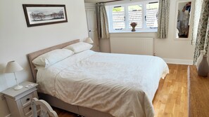 A neatly made bed in a room with a window, a bedside table with a lamp, and a framed picture on the wall.