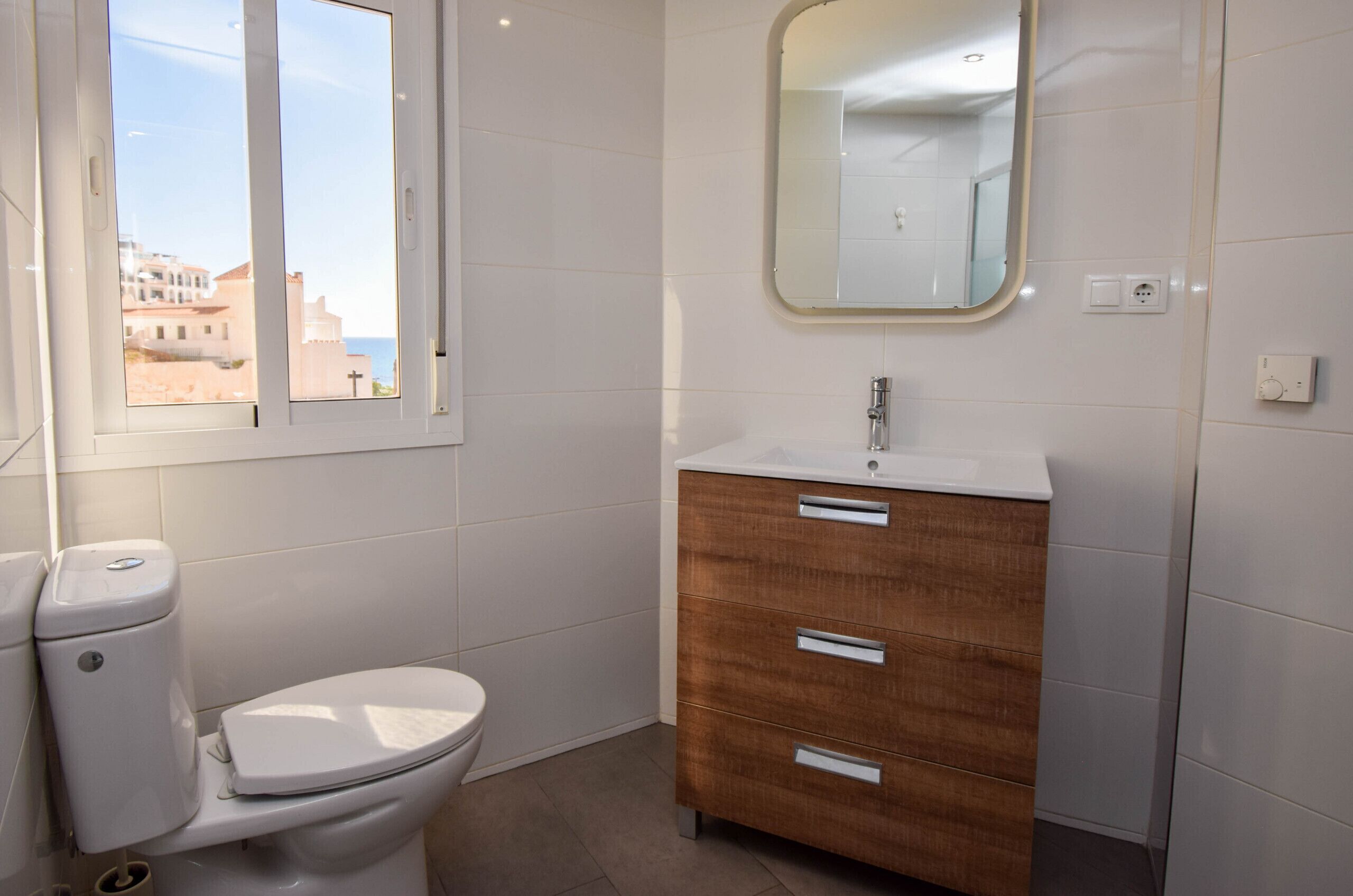 A bathroom with a toilet, a vanity with drawers, a mirror, and a window with a view of buildings.