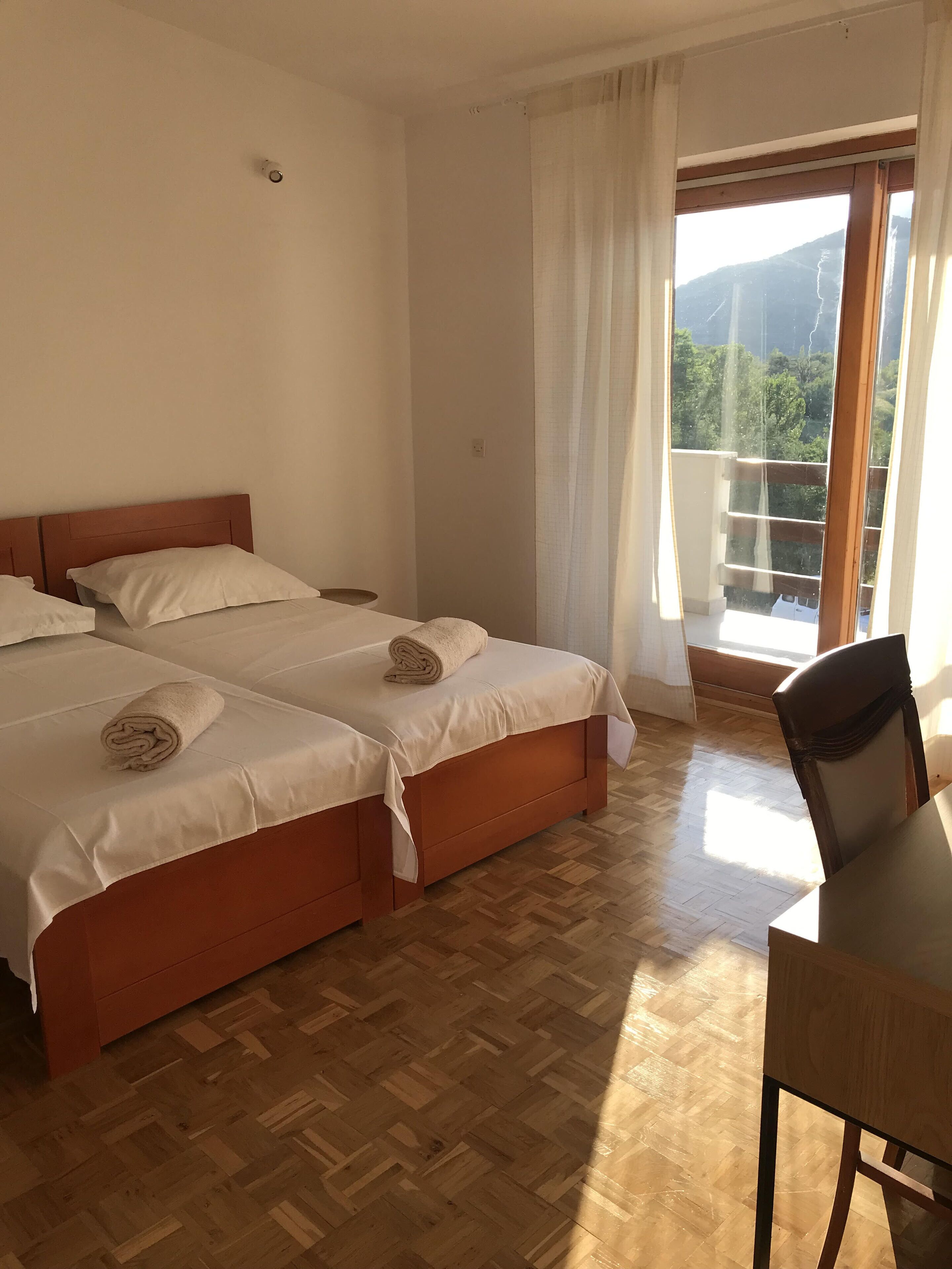 A hotel room with two beds, a wooden floor, a balcony with a view, and a table with a chair.
