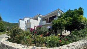 A modern two-story house with a stone retaining wall, surrounded by lush greenery and flowering plants. - Holiday house " Katy" on Bacina Lakes (PLOCE, PERACKO BLATO)