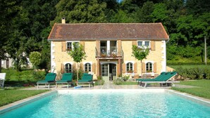 Une grande piscine entourée de chaises longues, un bâtiment en pierre avec un toit en tuiles et un jardin luxuriant.