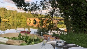 Une table dressée avec un saladier, des assiettes, des couverts et des verres, donnant sur un pont et un lac.