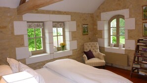 Une chambre avec un grand lit, deux fenêtres avec volets, une chaise et une bibliothèque.