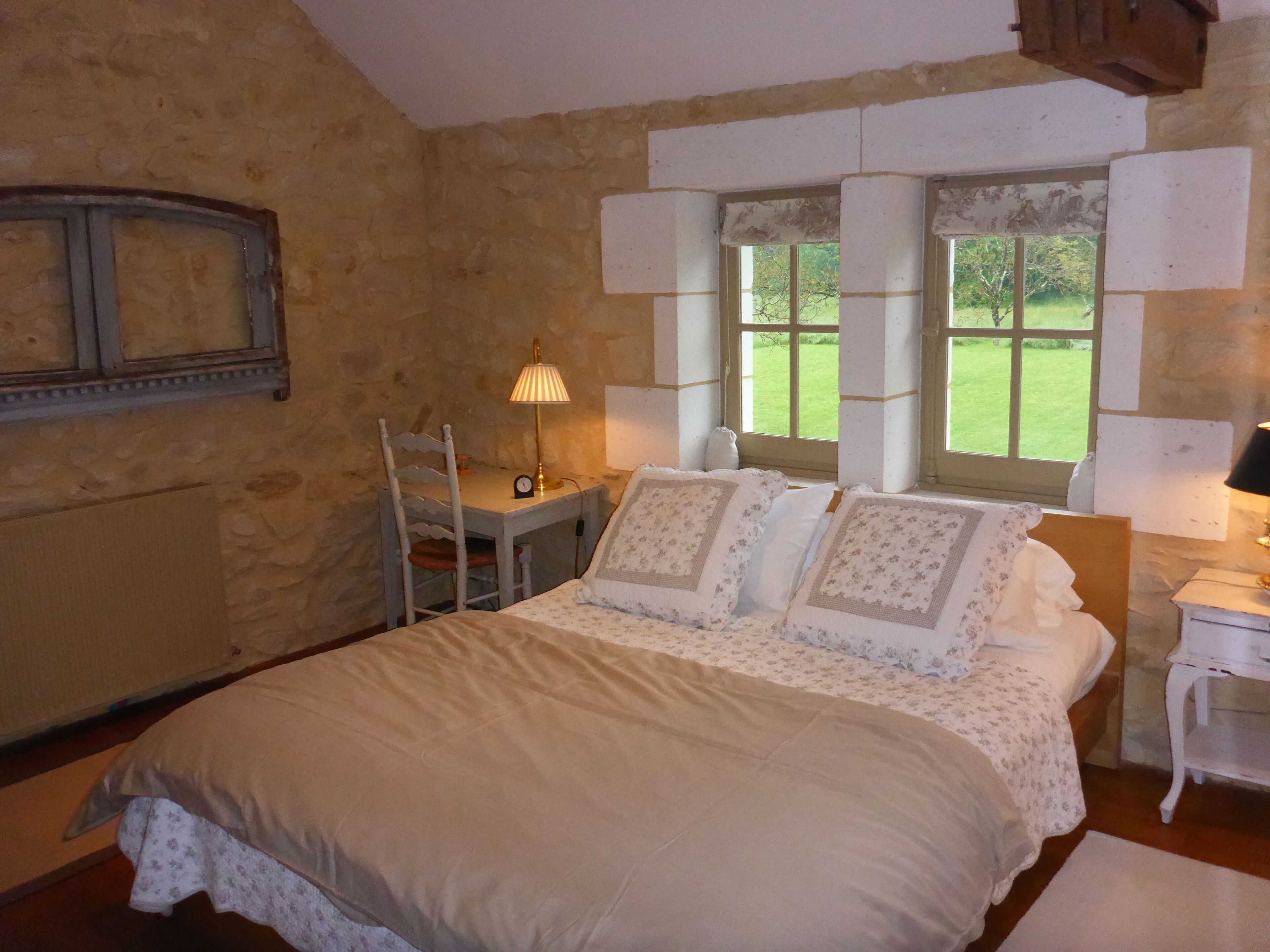A bedroom with a stone wall, a bed with floral bedding, a desk with a lamp, and a view of a green garden through large windows.