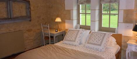 A bedroom with a stone wall, a bed with floral bedding, a desk with a lamp, and a view of a green garden through large windows.