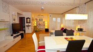 Un soggiorno moderno con una TV a schermo piatto, un tavolo da pranzo con sedie e un’accogliente zona salotto con divano e cuscini.