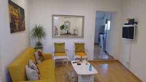 Ein Wohnzimmer mit einem gelben Sofa, zwei weißen Sesseln, einem Couchtisch und einem großen Spiegel. An der Wand befinden sich eine Zimmerpflanze und ein Gemälde.