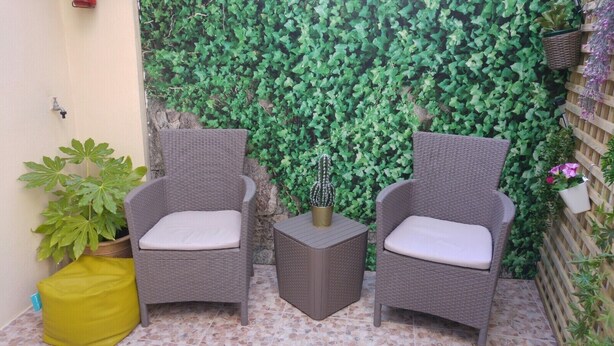 Zwei Korbstühle mit Kissen, ein kleiner runder Tisch und eine gelbe Tasche in einer Terrasseneinrichtung vor einer grünen, blätterreichen Wand.