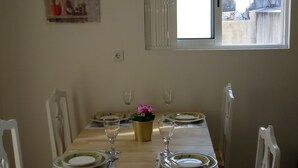 Ein Essbereich mit Tisch für vier Personen, gedeckt mit Tellern, Besteck und Weingläsern. Es gibt ein Fenster mit Jalousien, eine Steckdose und ein an der Wand hängendes Bild.