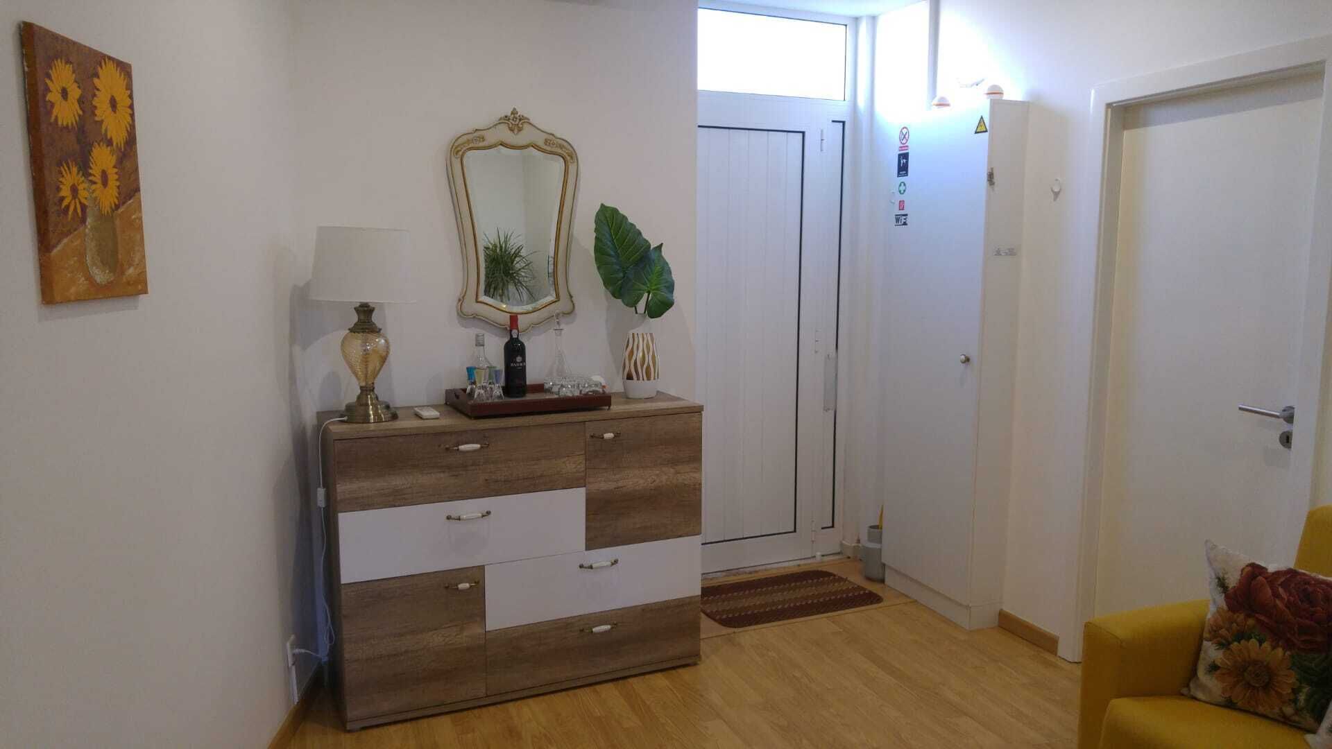 Ein Zimmer mit einem hölzernen Schminktisch, einem Spiegel, einer Lampe und einer Pflanze. Eine Tür und ein gelber Stuhl mit einem Blumenkissen sind ebenfalls vorhanden.