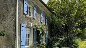 Une maison de campagne aux volets bleus, un jardin avec un petit étang et une pelouse agrémentée de deux bancs blancs.
