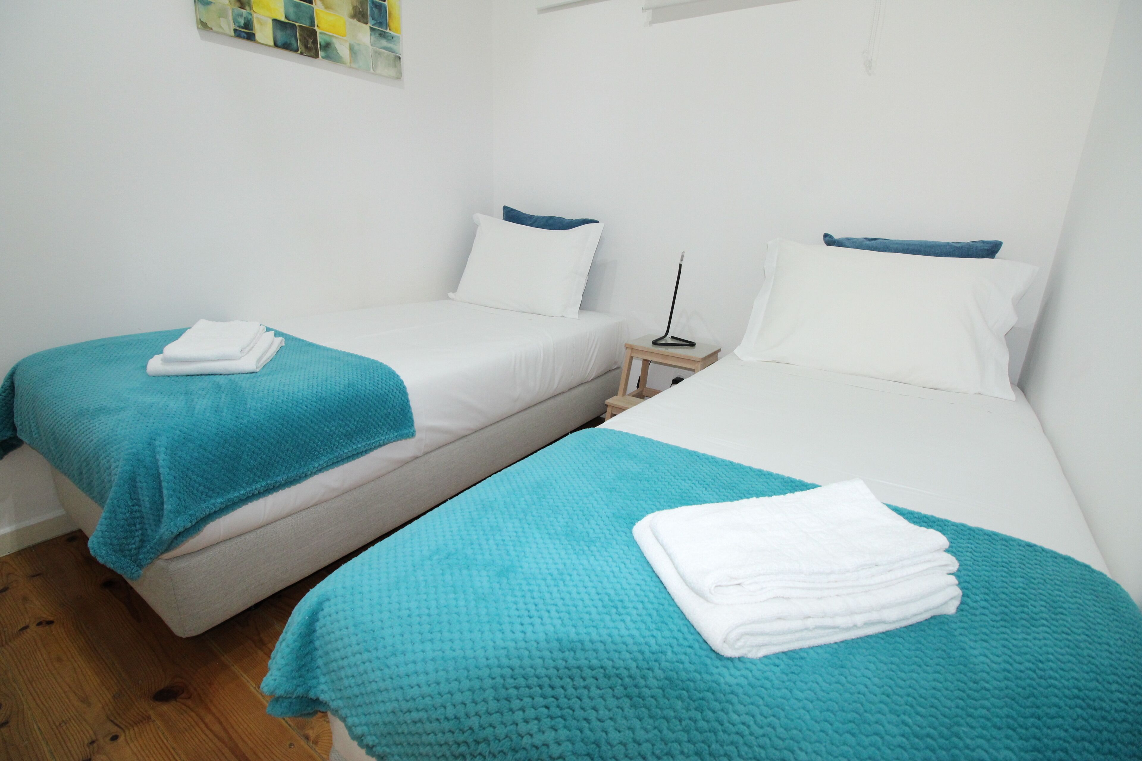 Habitación de hotel con dos camas individuales, cada una con una manta verde azulada y almohadas blancas. En cada cama hay una mesita de noche con una lámpara y una toalla blanca doblada.