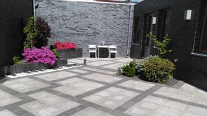 A courtyard with a tiled floor, two white tables, and chairs, surrounded by black brick walls and various plants.