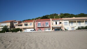 Una hilera de edificios de estilo mediterráneo con techos de tejas rojas, una playa arenosa en primer plano y un cielo azul despejado.
