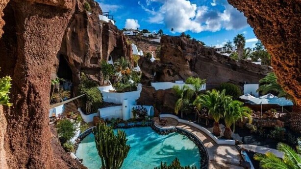 Un complexe hôtelier doté d’une piscine, entouré de bâtiments blancs et de palmiers, le tout se dressant sur un fond de falaises rocheuses et sous un ciel bleu azur.