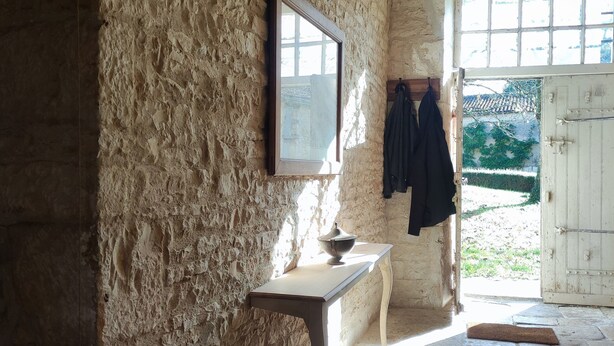 Une pièce rustique avec un mur en pierre, une table en bois, un manteau accroché et une fenêtre donnant sur un paysage verdoyant.