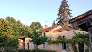 Un bâtiment en pierre, surmonté d’un toit en tuiles, entouré d’un jardin bien entretenu où l’on trouve divers arbustes et un petit arbre.