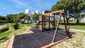 A playground with a swing set and a slide, surrounded by grass and trees, with a building in the background.
