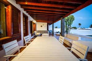 Un área de estar al aire libre con techo, vigas de madera, muebles blancos y vistas a la playa y palmeras.
