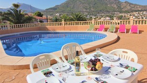 A poolside dining setup with a table set for two, a bottle of wine, and a fruit bowl.
