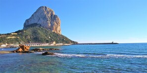 Paisagem costeira com uma formação rochosa imponente, águas azul-claras e um farol ao longe.