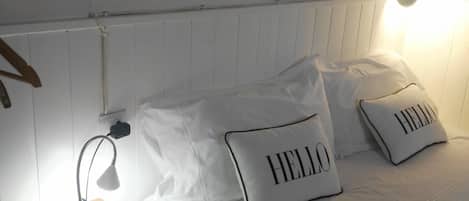 A neatly made bed with two pillows, one with "HELLO" on it, a bedside lamp, a wooden nightstand, and a wall-mounted light fixture.