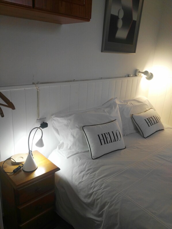 A neatly made bed with two pillows, one with "HELLO" on it, a bedside lamp, a wooden nightstand, and a wall-mounted light fixture.