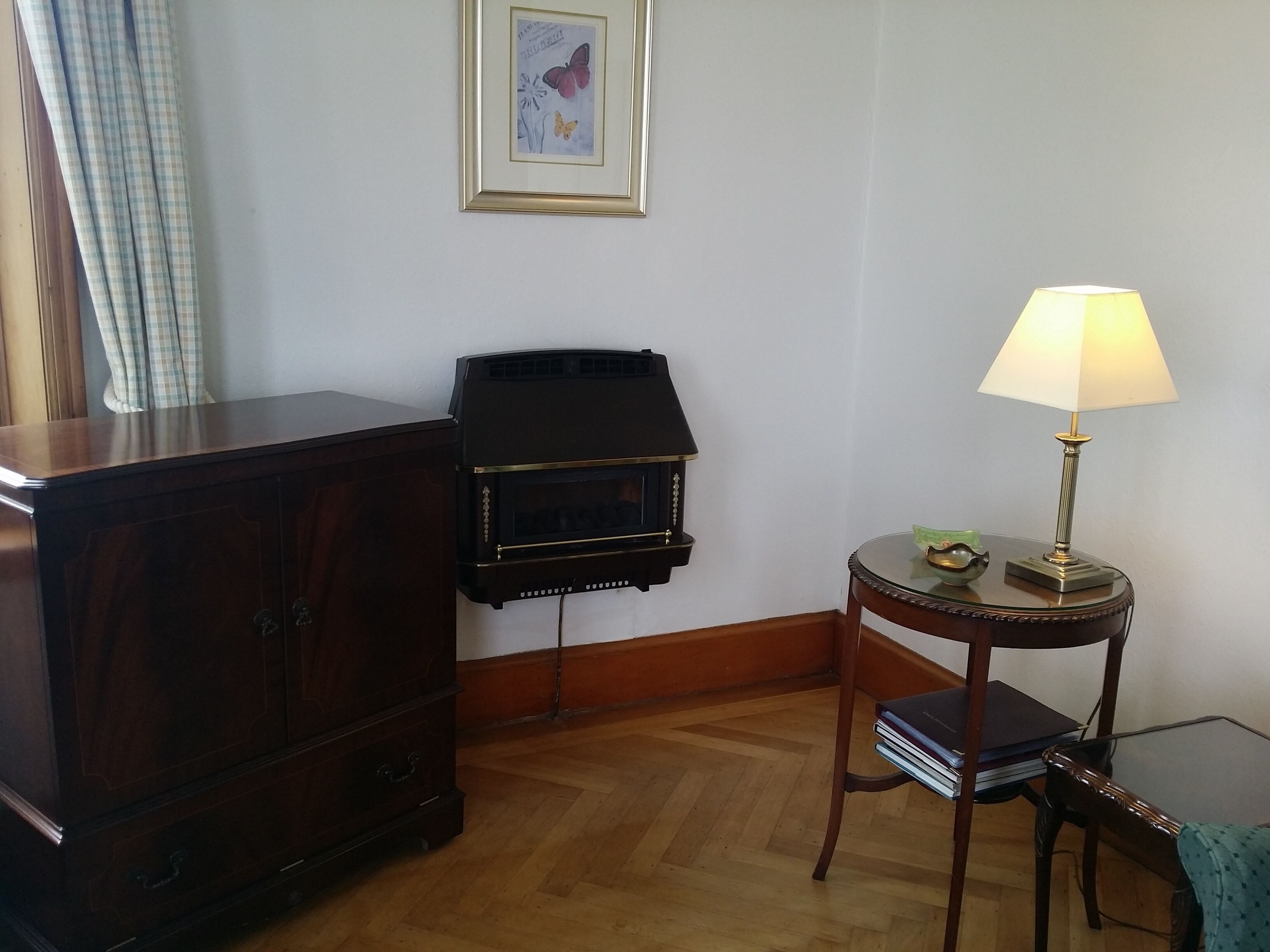 A room with a wooden cabinet, a black fireplace, a small round table with a lamp, and a chair.