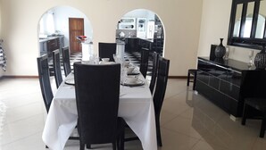 Un coin repas avec une longue table dressée pour six personnes, entourée de chaises. On aperçoit une cuisine à travers une arche en arrière-plan.
