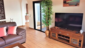 A living room with a brown leather sofa, a large flat-screen TV on a wooden stand, a tall bamboo plant, and a sliding glass door leading to an outdoor area. - Villa Anna - Beautiful exclusive Villa with pool  (Castillo Caleta de Fuste)