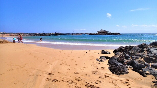 A beach with a rocky shoreline, a pier, and a building in the distance. - Villa Anna - Beautiful exclusive Villa with pool  (Castillo Caleta de Fuste)