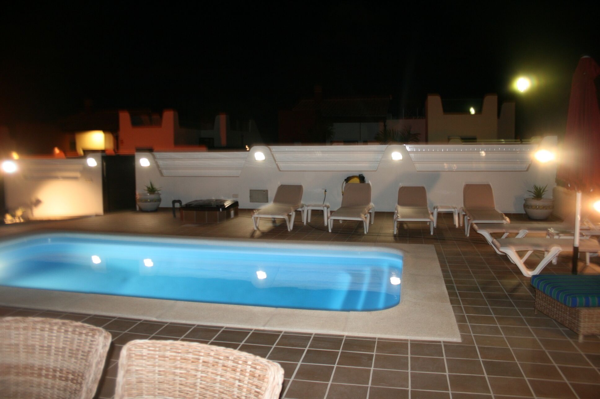 Une piscine entourée de chaises longues et de transats, ainsi que d’une terrasse carrelée et d’un petit muret agrémenté de luminaires.