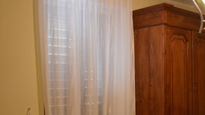 A bedroom with a wooden wardrobe, a bed with a red towel, and a window with sheer curtains.