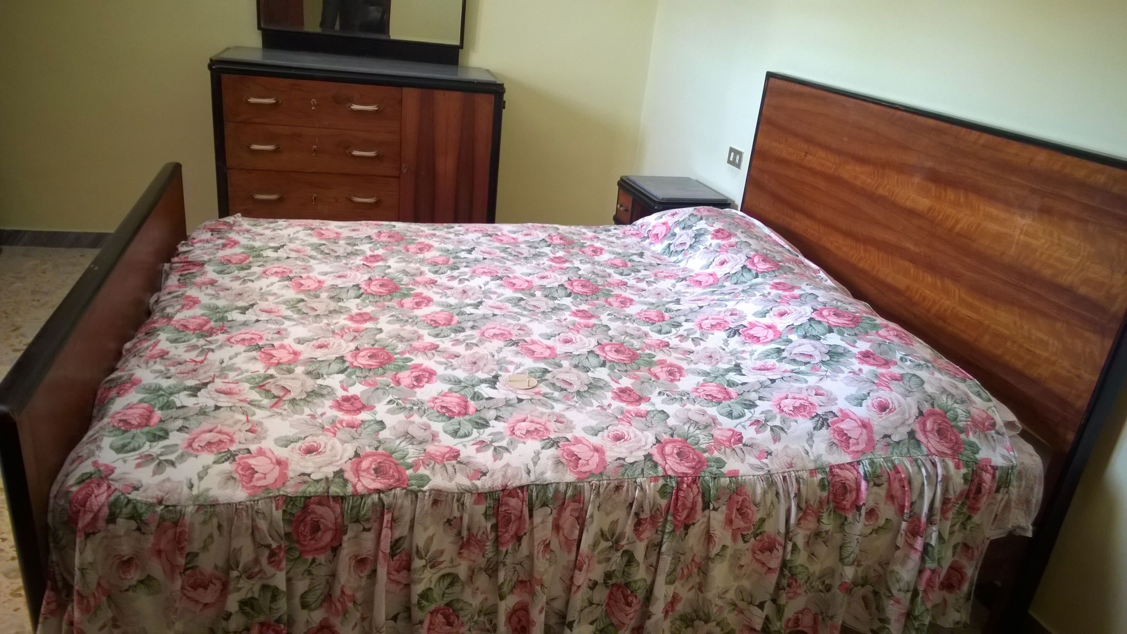 Een slaapkamer met een bloemenbedekker, een houten hoofdbord en een dressoir met lades.