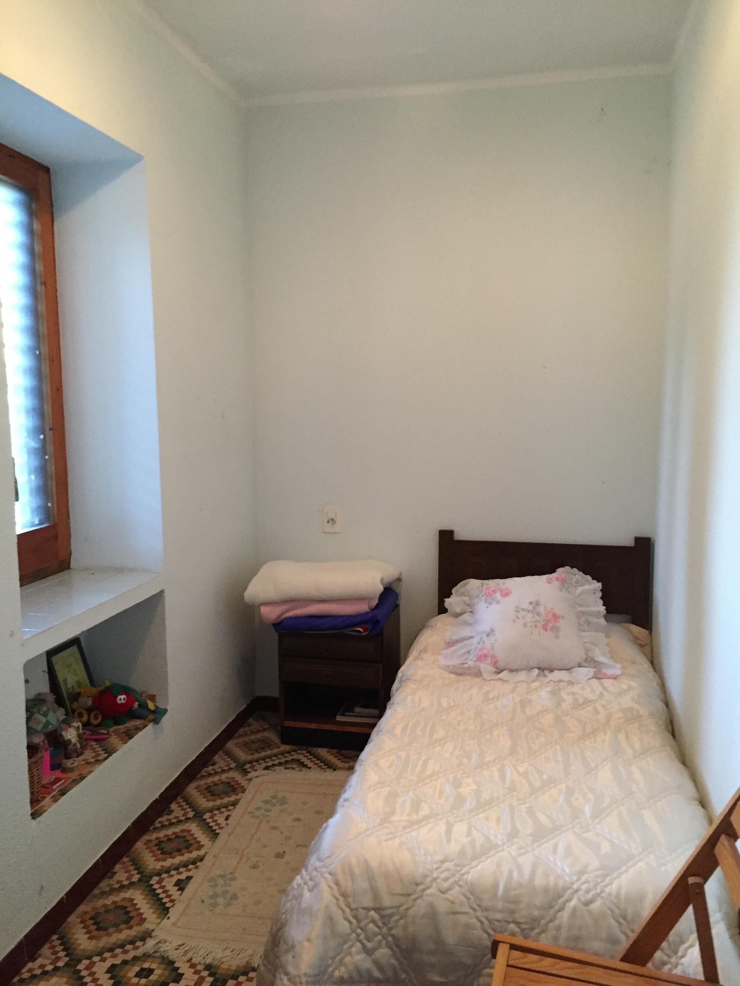 A small, neatly arranged bedroom with a single bed, a nightstand, a shelf with toys, and a patterned rug.