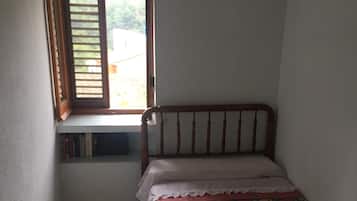 Una habitación pequeña y tenuemente iluminada con una cama individual, una ventana con contraventanas de madera y un suelo de baldosas.
