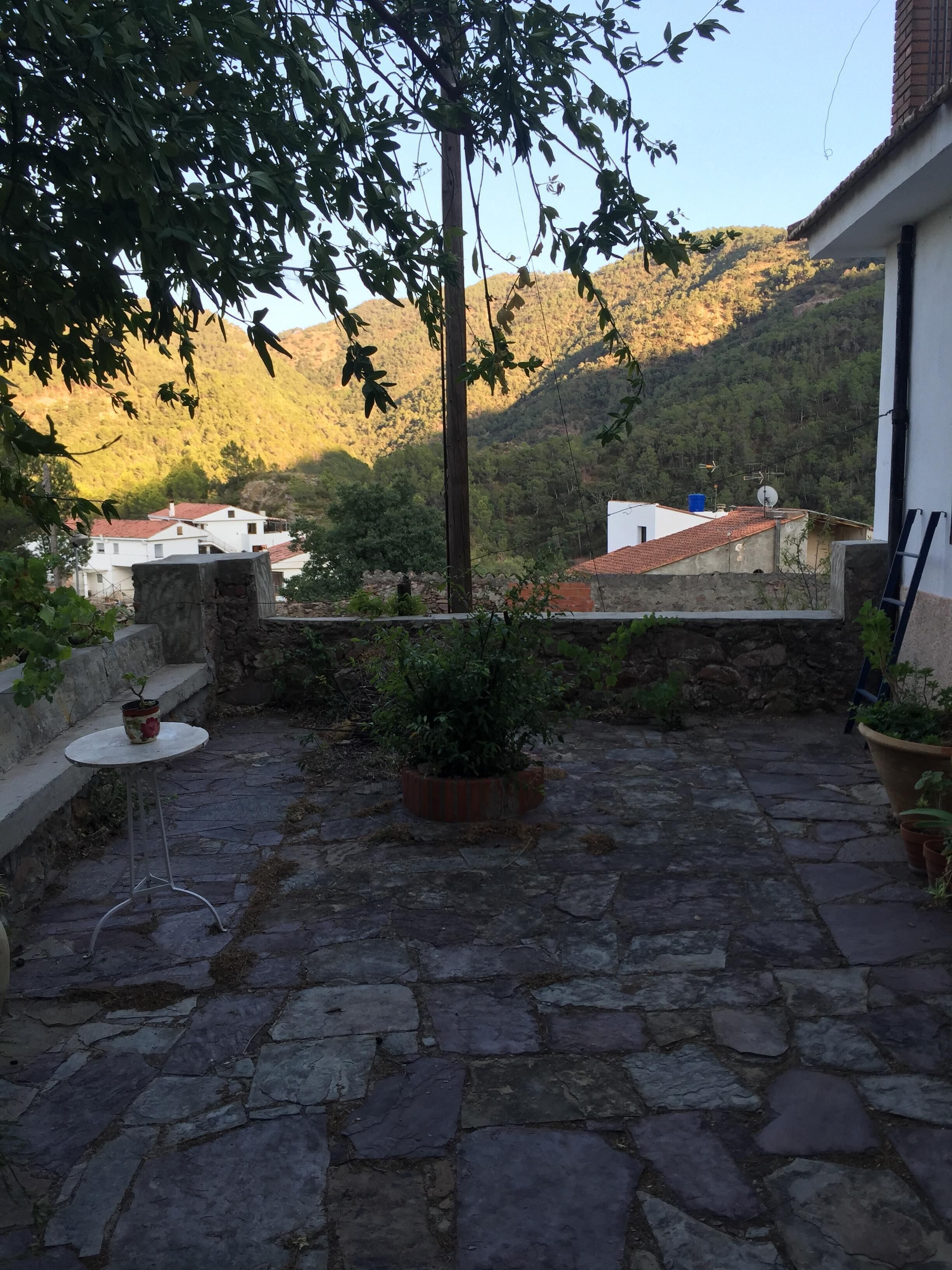 Un patio empedrado con una mesa pequeña y plantas en macetas, con vistas a una colina con edificios.