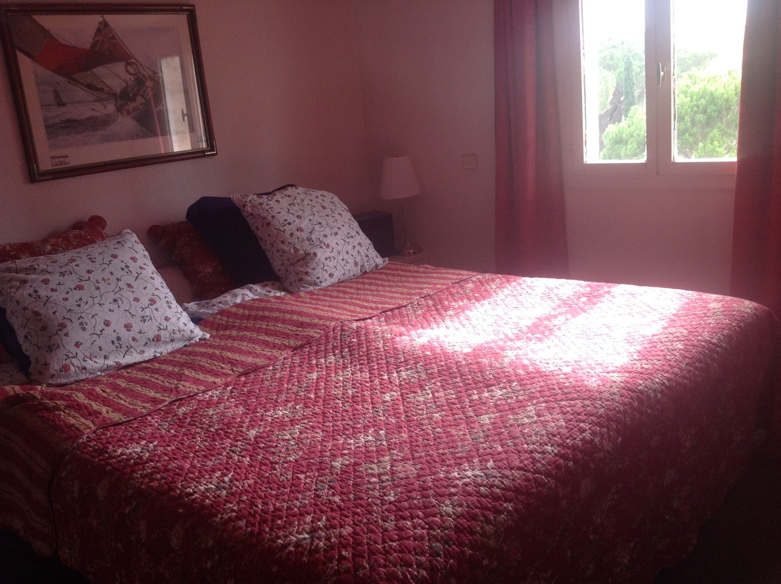 A bed with a red quilt, two pillows with floral patterns, and a framed picture on the wall.