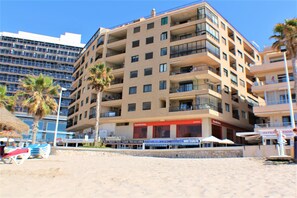 A multi-story hotel with balconies, located on a beach with palm trees and beach chairs.