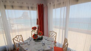 Ein Essbereich mit Tisch für zwei Personen, mit einer blumigen Tischdecke, einer Vase mit Sonnenblumen und Blick auf das Meer durch große Fenster.