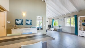 A spacious living area with a wooden dining table, white chairs, a kitchen area with white cabinets, and a large window with curtains.