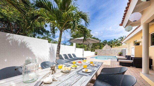 An outdoor dining area with a wooden table, black wicker chairs, a pool, and a white wall.
